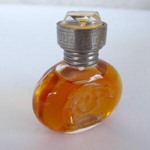 K&ouml;nnte beinhalten: Ein kleiner, ovaler Parf&uuml;mflakon aus Glas, gef&uuml;llt mit einer bernsteinfarbenen Fl&uuml;ssigkeit. Der Flakon hat einen transparenten, facettierten Verschluss und eine silberfarbene Metallkappe mit griechischem M&auml;ander und Medusenhaupt.