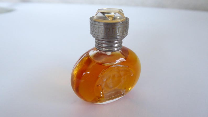 K&ouml;nnte beinhalten: Ein kleiner, ovaler Parf&uuml;mflakon aus Glas, gef&uuml;llt mit einer bernsteinfarbenen Fl&uuml;ssigkeit. Der Flakon hat einen transparenten, facettierten Verschluss und eine silberfarbene Metallkappe mit griechischem M&auml;ander und Medusenhaupt.