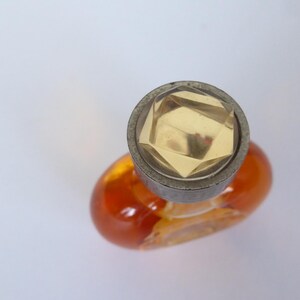 K&ouml;nnte beinhalten: Ein kleiner, ovaler Parf&uuml;mflakon aus Glas, gef&uuml;llt mit einer bernsteinfarbenen Fl&uuml;ssigkeit. Der Flakon hat einen runden, metallisch wirkenden Verschluss mit einem facettierten, klaren, geometrischen kristallartigen Einsatz in der Mitte.