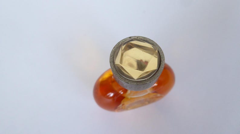 K&ouml;nnte beinhalten: Ein kleiner, ovaler Parf&uuml;mflakon aus Glas, gef&uuml;llt mit einer bernsteinfarbenen Fl&uuml;ssigkeit. Der Flakon hat einen runden, metallisch wirkenden Verschluss mit einem facettierten, klaren, geometrischen kristallartigen Einsatz in der Mitte.