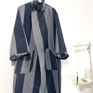 May include: A long, striped, black and grey coat with a high collar and pockets. The coat is hanging on a hanger.