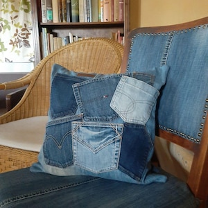 Op de afbeelding: Een blauw denim kussen met meerdere zakken die aan elkaar zijn genaaid. Het kussen ligt op een blauwe denim stoel met een houten frame.