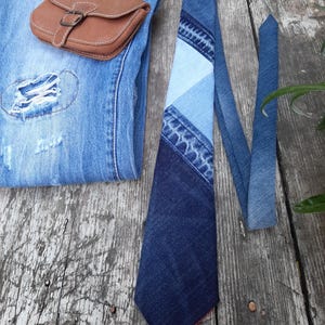 Può includere: Una cravatta in denim con un design patchwork in varie tonalità di blu, con fodera rossa. Una piccola pochette in pelle marrone con fibbia è appoggiata su un paio di jeans blu con una toppa strappata.