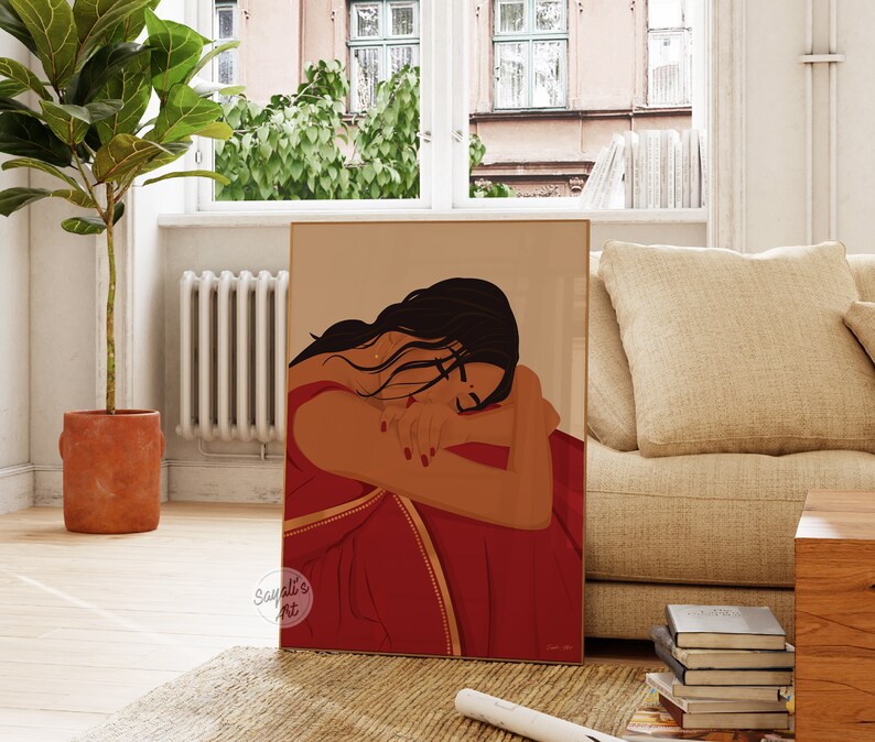 Puede incluir: Una ilustraci&oacute;n digital de una mujer con cabello largo y oscuro, vestida con un sari rojo y dorado, con la cabeza apoyada en el brazo. La ilustraci&oacute;n est&aacute; enmarcada y se exhibe en una pared de una sala de estar.
