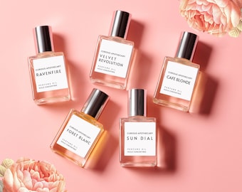 May include: Five small glass bottles of perfume oil on a pink background. The bottles are labelled with the brand name "Curious Apothecary" and the names of the scents: "Ravenfire", "Velvet Revolution", "Cafe Blonde", "Foret Blanc", and "Sun Dial".