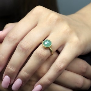 May include: A gold ring with a round, green gemstone. The ring is on a person's finger.