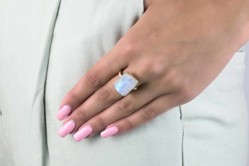 May include: A gold ring with a large, rectangular, iridescent moonstone. The ring is on a person's finger.