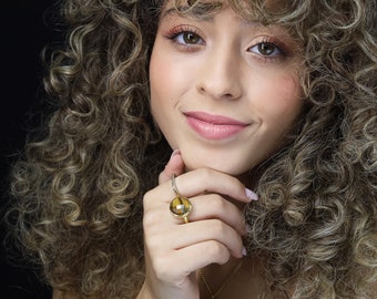 Op de afbeelding: Een vrouw met krullend bruin haar draagt een gouden ring met een grote, ronde, gele edelsteen. De ring zit aan haar rechterhand en ze houdt haar kin vast met haar linkerhand.