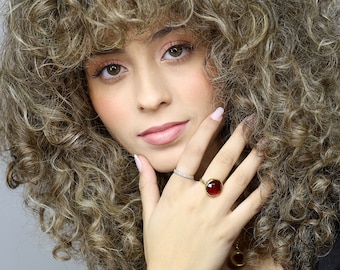 May include: A woman with curly brown hair is wearing a gold ring with a large red gemstone. The ring is on her right hand, and she is holding her hand up to her face.
