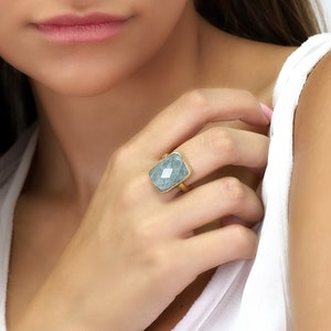 May include: A gold ring with a large, rectangular, light blue gemstone. The ring is on a person's finger.