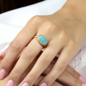 Puede incluir: Un anillo de oro con una piedra ovalada de color azul claro. El anillo est&aacute; en el dedo de una persona.
