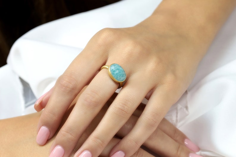 Puede incluir: Un anillo de oro con una piedra ovalada de color azul claro. El anillo est&aacute; en el dedo de una persona.