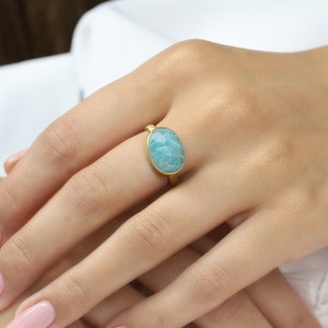 Puede incluir: Un anillo de oro con una piedra ovalada de color azul claro. El anillo est&aacute; en el dedo de una persona.