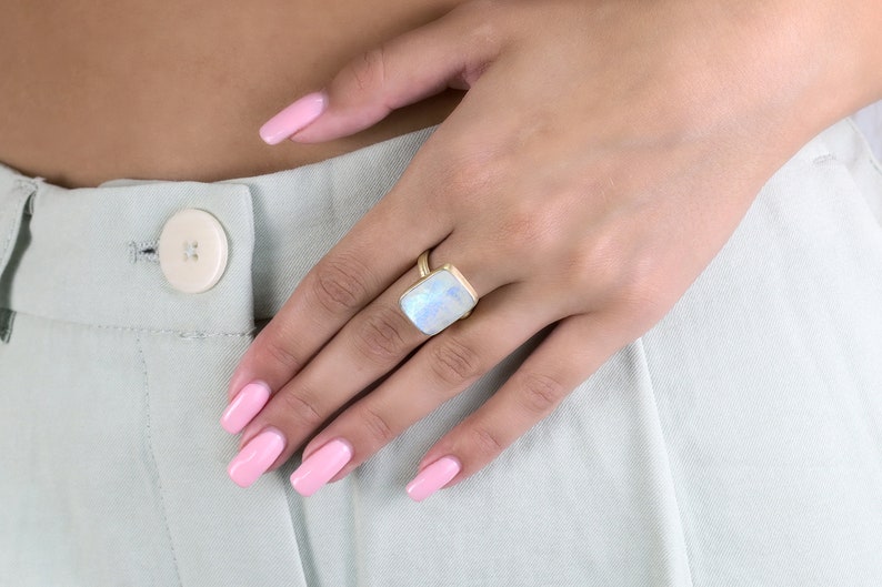 May include: A gold ring with a large, rectangular, iridescent moonstone. The ring is on a person's finger.