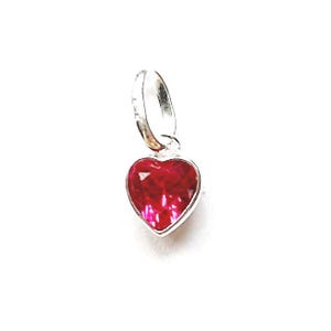 May include: A heart-shaped pendant featuring a bright red gemstone, set within a silver-coloured metal frame. The pendant hangs from a small loop, suitable for attaching to a necklace or bracelet. The gemstone's surface is faceted, reflecting light.