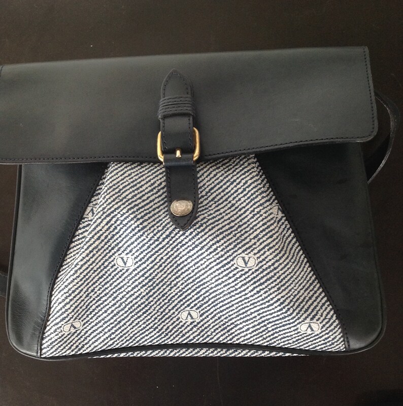 May include: A blue and white patterned shoulder bag with a black leather flap and a gold buckle. The bag has a strap and a small logo on the front.