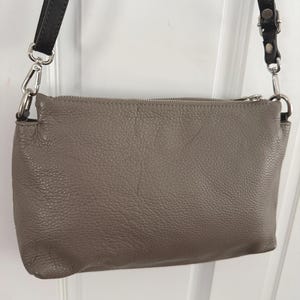 May include: A taupe leather crossbody bag with a textured finish. It features a silver zip and black leather straps. The straps are connected to the bag with silver metal clasps.