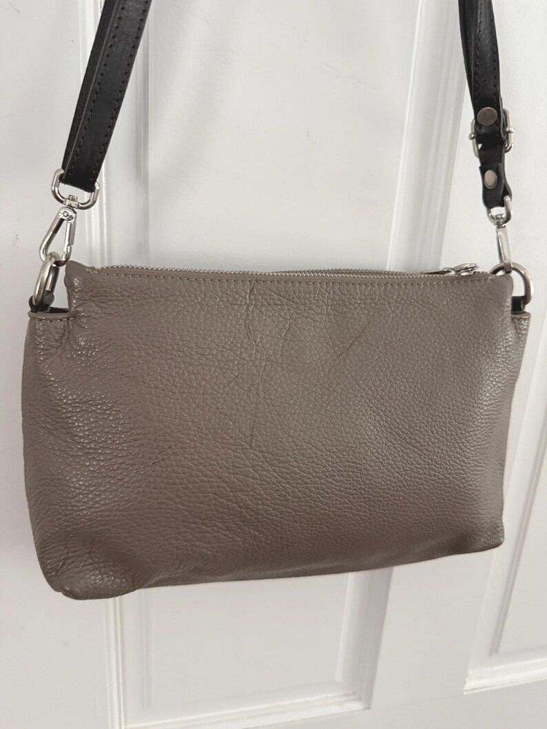 May include: A taupe leather crossbody bag with a textured finish. It features a silver zip and black leather straps. The straps are connected to the bag with silver metal clasps.