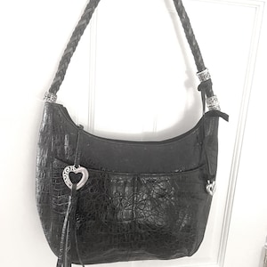 Puede incluir: Bolso de hombro de cuero negro con textura de cocodrilo. El bolso presenta una correa trenzada, herrajes plateados y un dije en forma de coraz&oacute;n con borla. El bolso cuelga contra una puerta blanca.