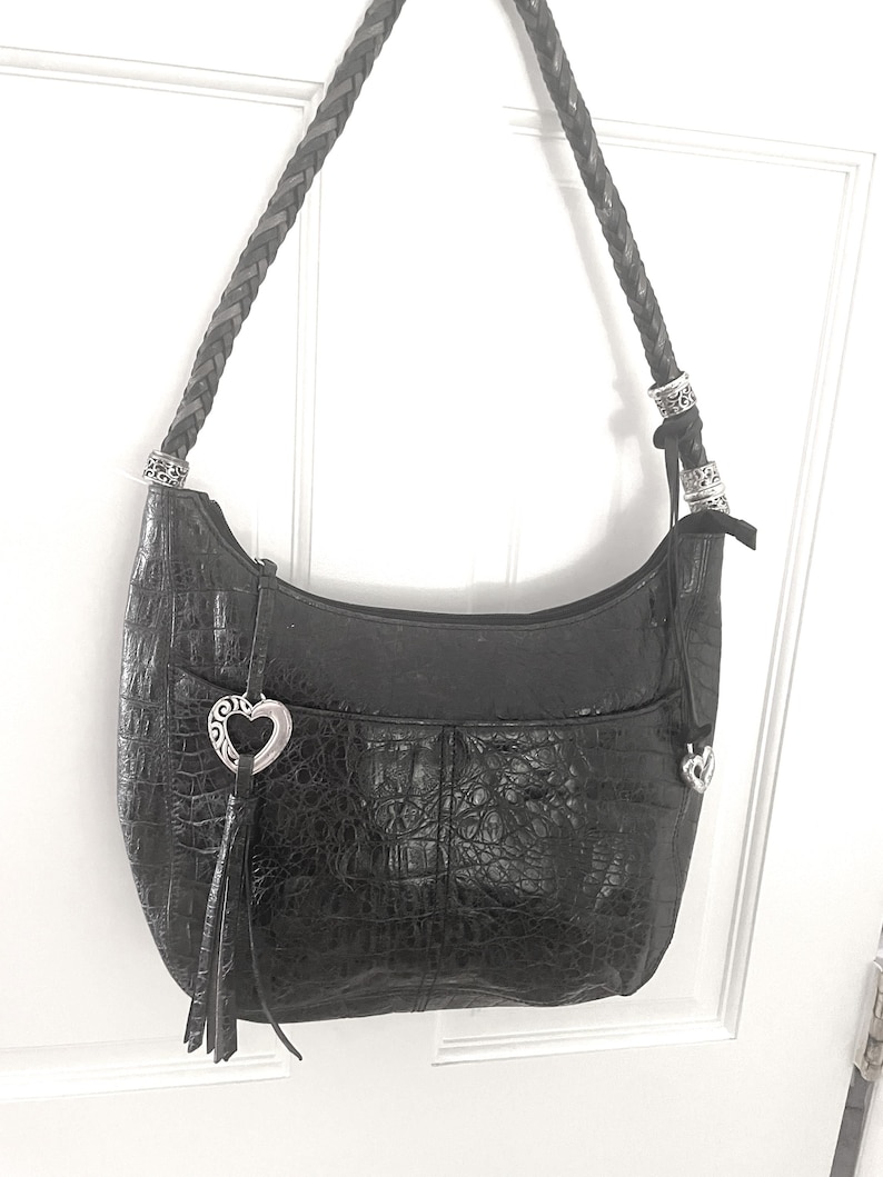 Puede incluir: Bolso de hombro de cuero negro con textura de cocodrilo. El bolso presenta una correa trenzada, herrajes plateados y un dije en forma de coraz&oacute;n con borla. El bolso cuelga contra una puerta blanca.