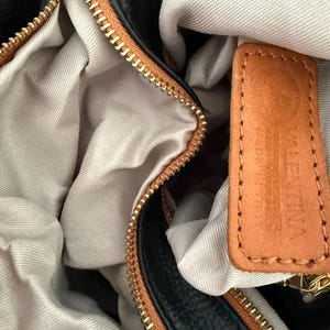 May include: Close-up of a black and brown leather handbag with a gold zipper. The interior is a light beige fabric. A small brown leather tag is visible.