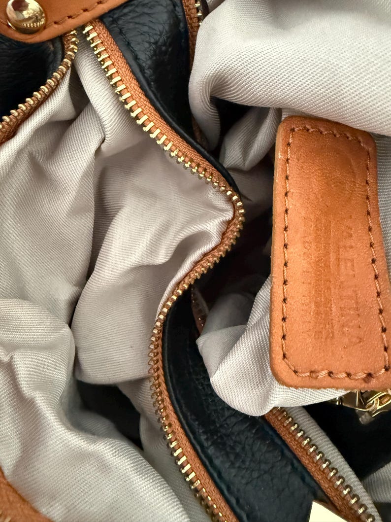 May include: Close-up of a black and brown leather handbag with a gold zipper. The interior is a light beige fabric. A small brown leather tag is visible.