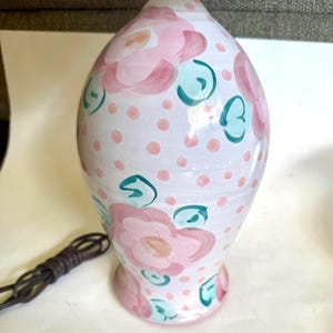 May include: A white ceramic lamp with a floral design. The lamp features pink flowers, teal leaves, and pink polka dots. A brown electrical cord is visible. The lamp has a bulb base and a gold-colored top.