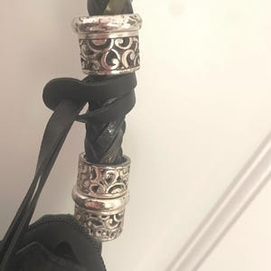 Puede incluir: Primer plano de una correa de bolso de cuero negro con un dise&ntilde;o trenzado. La correa presenta dos anillos decorativos plateados con un patr&oacute;n ornamental y arremolinado. El cuerpo del bolso es visible en la parte inferior, tambi&eacute;n en cuero negro.