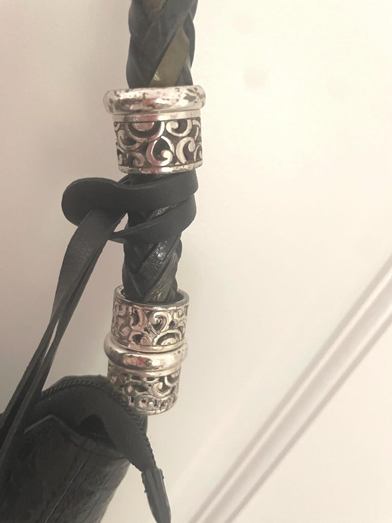 Puede incluir: Primer plano de una correa de bolso de cuero negro con un dise&ntilde;o trenzado. La correa presenta dos anillos decorativos plateados con un patr&oacute;n ornamental y arremolinado. El cuerpo del bolso es visible en la parte inferior, tambi&eacute;n en cuero negro.
