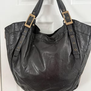 May include: A large black slouchy tote bag with a textured finish. The bag features two shoulder straps attached with rectangular brass-toned buckles and adjustable strap detailing with small circular grommets on the front panels.