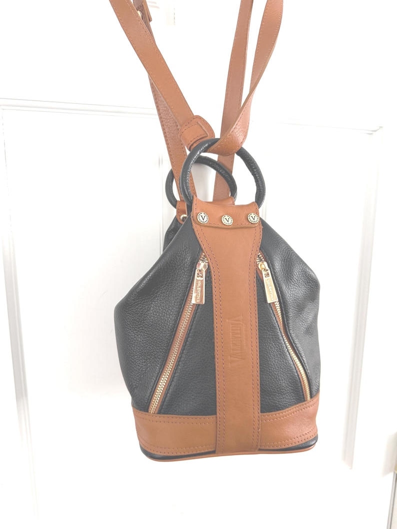 May include: Black and brown leather backpack with gold zippers and accents. The backpack has a rounded shape with a brown base and straps. The word "Valentina" is printed vertically on the front of the bag.