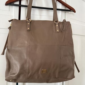 May include: Taupe leather tote bag with double handles and a zipper closure. The bag has a rectangular shape with a stitched design and gold-tone hardware. A small gold logo is visible on the front.