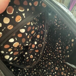 Puede incluir: Vista interior de un bolso negro con forro estampado. El forro presenta un patr&oacute;n repetido de formas marrones, beige y blancas sobre un fondo oscuro. La cremallera y las costuras del bolso son visibles.