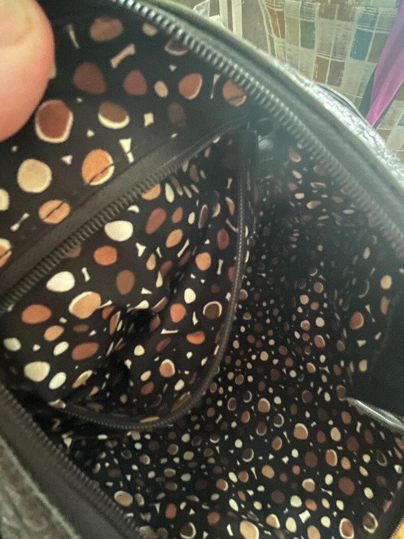 Puede incluir: Vista interior de un bolso negro con forro estampado. El forro presenta un patr&oacute;n repetido de formas marrones, beige y blancas sobre un fondo oscuro. La cremallera y las costuras del bolso son visibles.
