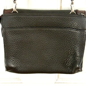 May include: A dark brown leather crossbody bag with a textured surface. The bag has a rectangular shape with a flap closure and a long, adjustable strap. Silver-tone hardware and brown accents are visible.