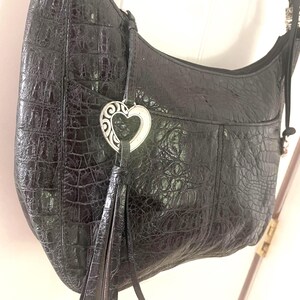 Puede incluir: Bolso de hombro de cuero negro con textura de cocodrilo. El bolso tiene un charm plateado en forma de coraz&oacute;n y una borla. La correa negra tiene detalles plateados. Tiene un bolsillo frontal.