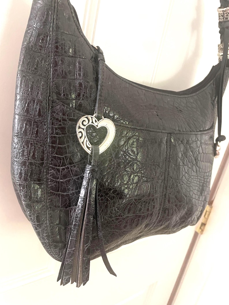 Puede incluir: Bolso de hombro de cuero negro con textura de cocodrilo. El bolso tiene un charm plateado en forma de coraz&oacute;n y una borla. La correa negra tiene detalles plateados. Tiene un bolsillo frontal.