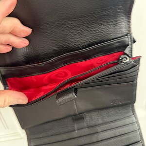 May include: Black leather wallet with a red interior. The wallet has multiple compartments, including card slots and a zippered section. The interior features a decorative pattern. The wallet is open, displaying its contents.