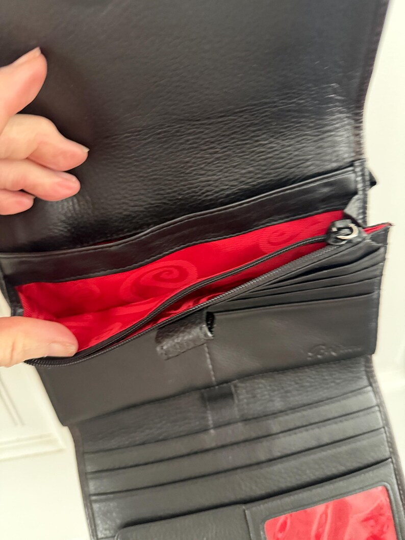 May include: Black leather wallet with a red interior. The wallet has multiple compartments, including card slots and a zippered section. The interior features a decorative pattern. The wallet is open, displaying its contents.