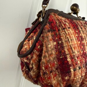 K&ouml;nnte beinhalten: Eine strukturierte Handtasche mit einem gewebten Muster in Rot-, Orange- und Beiget&ouml;nen. Die Tasche hat einen dunklen braunen Lederbesatz, einen goldfarbenen Kettenriemen und einen Verschluss. Das Design der Tasche ist von Vintage-Stil inspiriert.