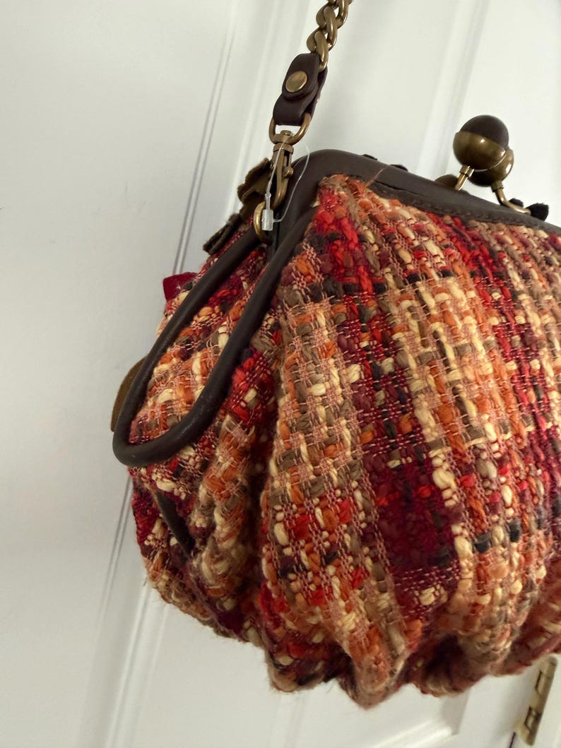 K&ouml;nnte beinhalten: Eine strukturierte Handtasche mit einem gewebten Muster in Rot-, Orange- und Beiget&ouml;nen. Die Tasche hat einen dunklen braunen Lederbesatz, einen goldfarbenen Kettenriemen und einen Verschluss. Das Design der Tasche ist von Vintage-Stil inspiriert.