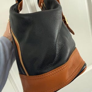 May include: Black and brown leather handbag with gold-tone hardware. The bag features a black body with a brown base and accents. A gold zipper runs down the side. The bag has a top handle and a shoulder strap.