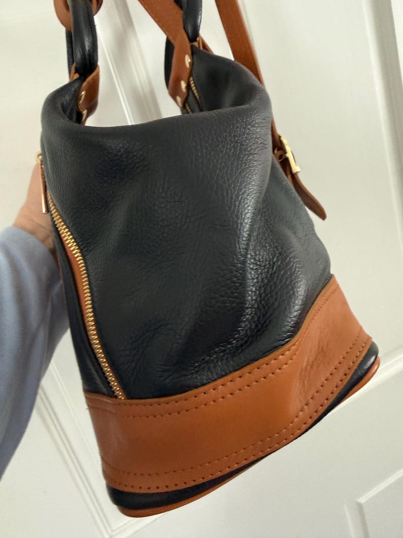 May include: Black and brown leather handbag with gold-tone hardware. The bag features a black body with a brown base and accents. A gold zipper runs down the side. The bag has a top handle and a shoulder strap.