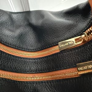 May include: Close-up of a black leather handbag with a textured surface. The bag features two gold-colored zippers with brown trim and the name "VALENTINA" embossed on the zipper pulls. The bag's design is simple and elegant.