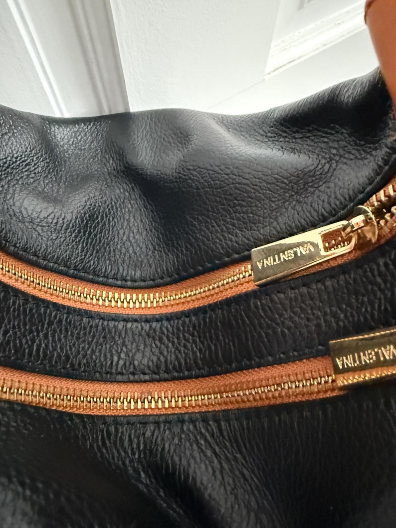 May include: Close-up of a black leather handbag with a textured surface. The bag features two gold-colored zippers with brown trim and the name "VALENTINA" embossed on the zipper pulls. The bag's design is simple and elegant.