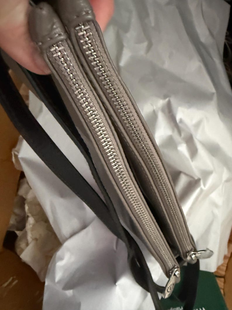 May include: Two grey zippered pouches with silver zips and black straps. The pouches are made of a smooth material and have a simple, minimalist design. The zips are closed, and the pouches appear to be empty. The background is a crumpled white surface.