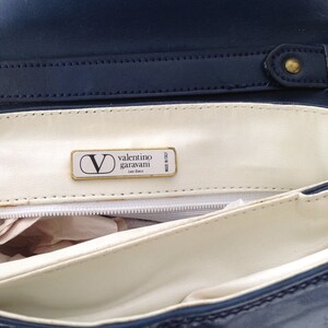 May include: Inside of a blue and white striped handbag with a white leather lining. The lining has a gold label that reads "valentino garavani Les Sacs Made in Italy."