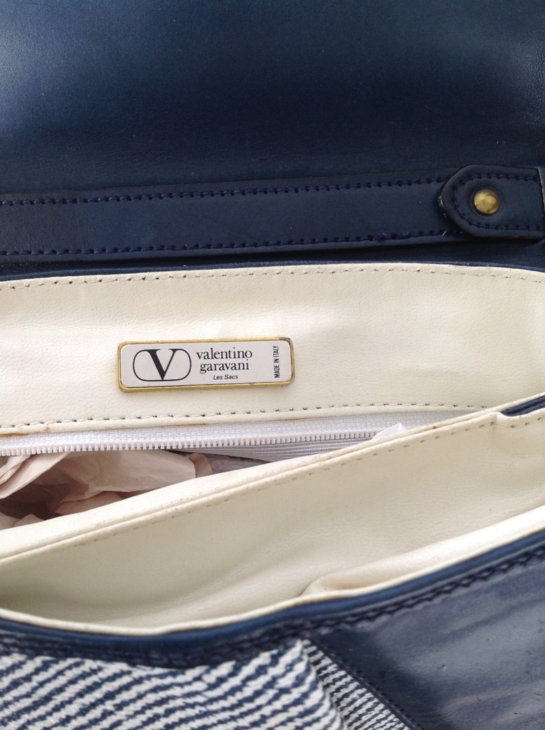May include: Inside of a blue and white striped handbag with a white leather lining. The lining has a gold label that reads "valentino garavani Les Sacs Made in Italy."