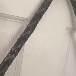 Puede incluir: Primer plano de una correa de cuero trenzado en tonos marr&oacute;n oscuro y negro. La trenza, probablemente para un bolso o accesorio, est&aacute; finamente tejida, mostrando un patr&oacute;n texturizado. Cuelga sobre un fondo blanco.