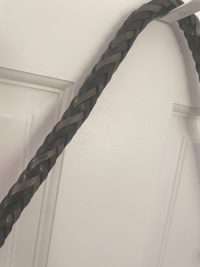 Puede incluir: Primer plano de una correa de cuero trenzado en tonos marr&oacute;n oscuro y negro. La trenza, probablemente para un bolso o accesorio, est&aacute; finamente tejida, mostrando un patr&oacute;n texturizado. Cuelga sobre un fondo blanco.